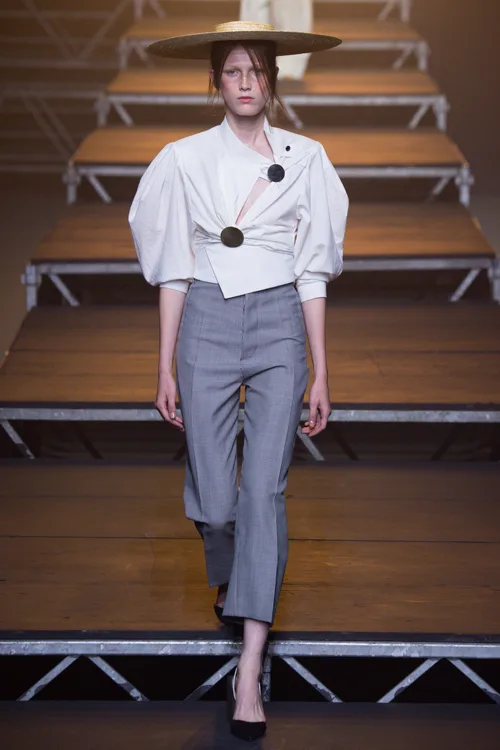 Simon Porte Jacquemus, người mẫu mặc áo trắng tay phồng, quần tây cropped màu xám và đội mũ.