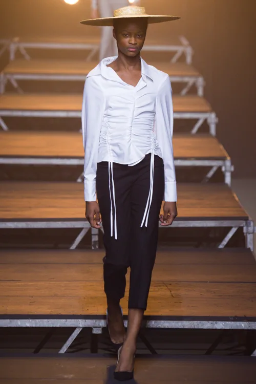 Simon Porte Jacquemus: người mẫu đội mũ, mặc áo sơ mi ôm màu trắng có 2 hàng dây cột và quần cropped màu đen.