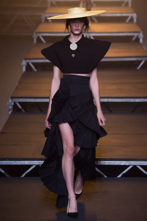 Simon Porte Jacquemus: người mẫu mặc áo crop top đen, chân váy xòe nhiều lớp màu đen và mũ cói.